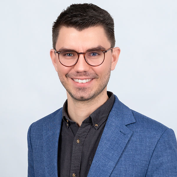 Ein lächelnder Mann mit Brille in einem dunklen Hemd und blauen Sakko.