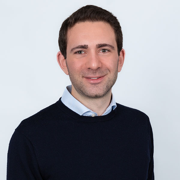 Ein Mann in einem dunkelblauen Pullover und einem hellen Hemd lächelt in die Kamera.