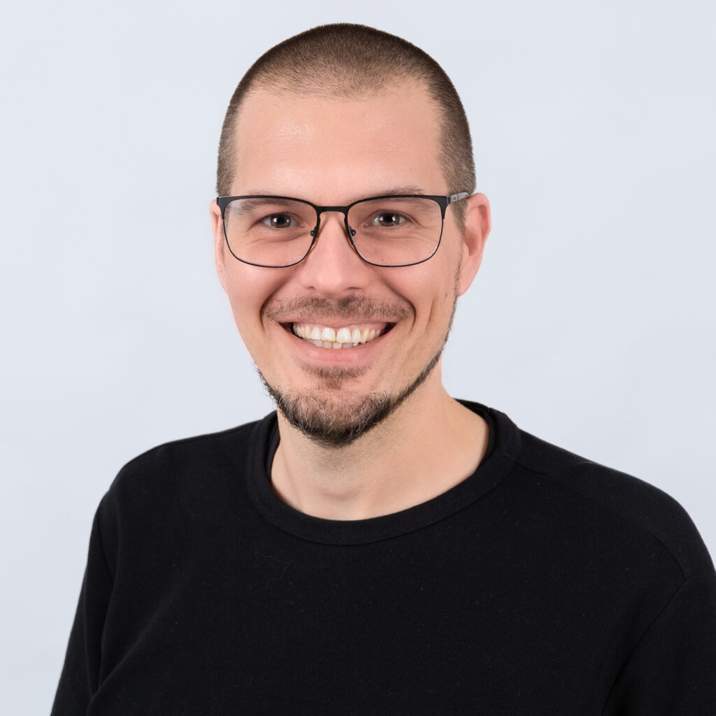 Porträt eines jungen Mannes mit Brille und Bart, freundlich lächelnd.