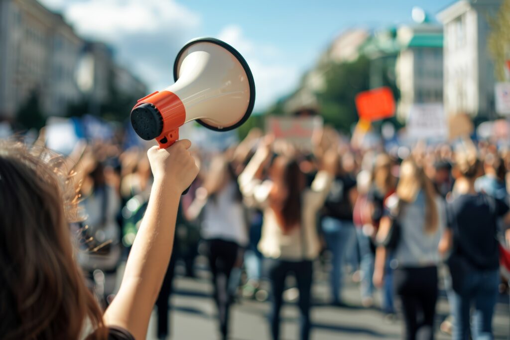Frau hält Megafon, Menschen demonstrieren im Hintergrund.